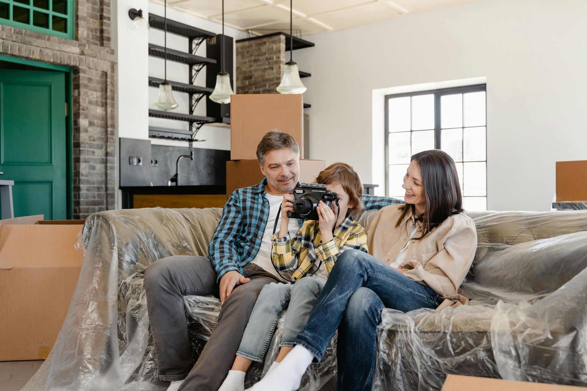 Family sits on a couch, child holding camera, boxes in new home.