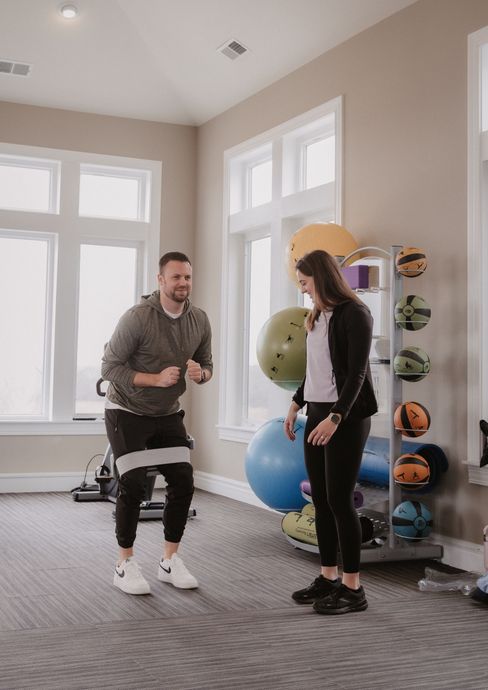 A person in gym clothes performs a band-resisted squat while a trainer observes in a brightly lit fitness studio.
