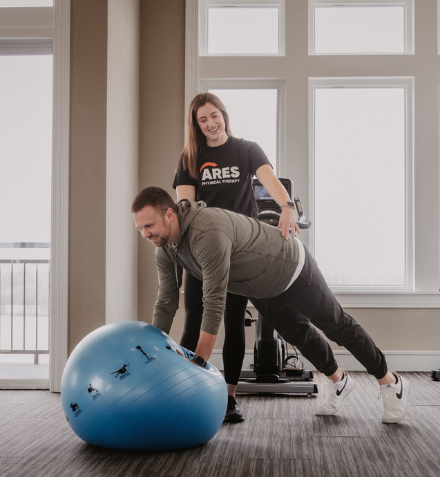 A fitness professional assists a man in a plank exercise with his hands balanced on a blue stability ball.