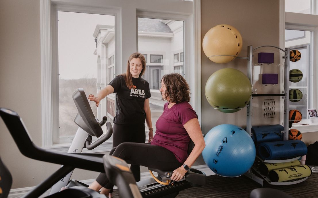 A fitness trainer in a black shirt helps a client use an exercise machine in a bright gym with exercise balls.