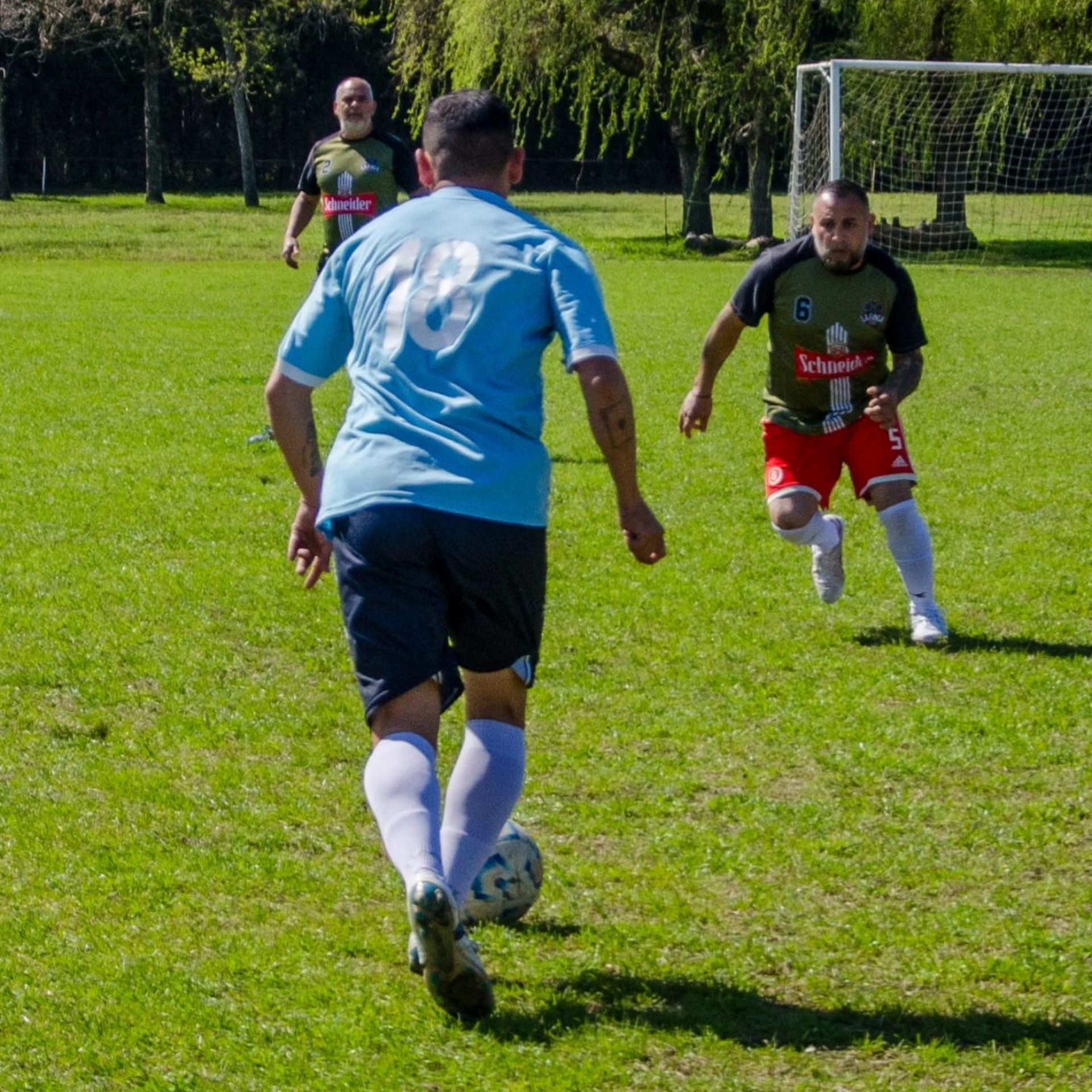 Un jugador de fútbol con una camiseta azul dribla un balón en un campo de césped mientras dos oponentes se acercan.