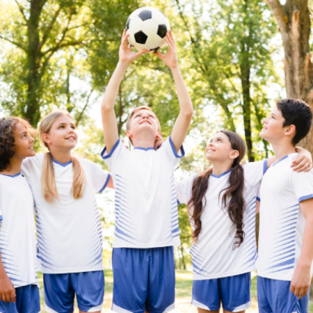 Cinco niños con uniformes de fútbol blancos y azules están parados en un parque, mirando hacia arriba a un balón de fútbol