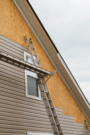 A metal ladder scaffold set up against the side of a house, where siding is partially installed over plywood sheathing.