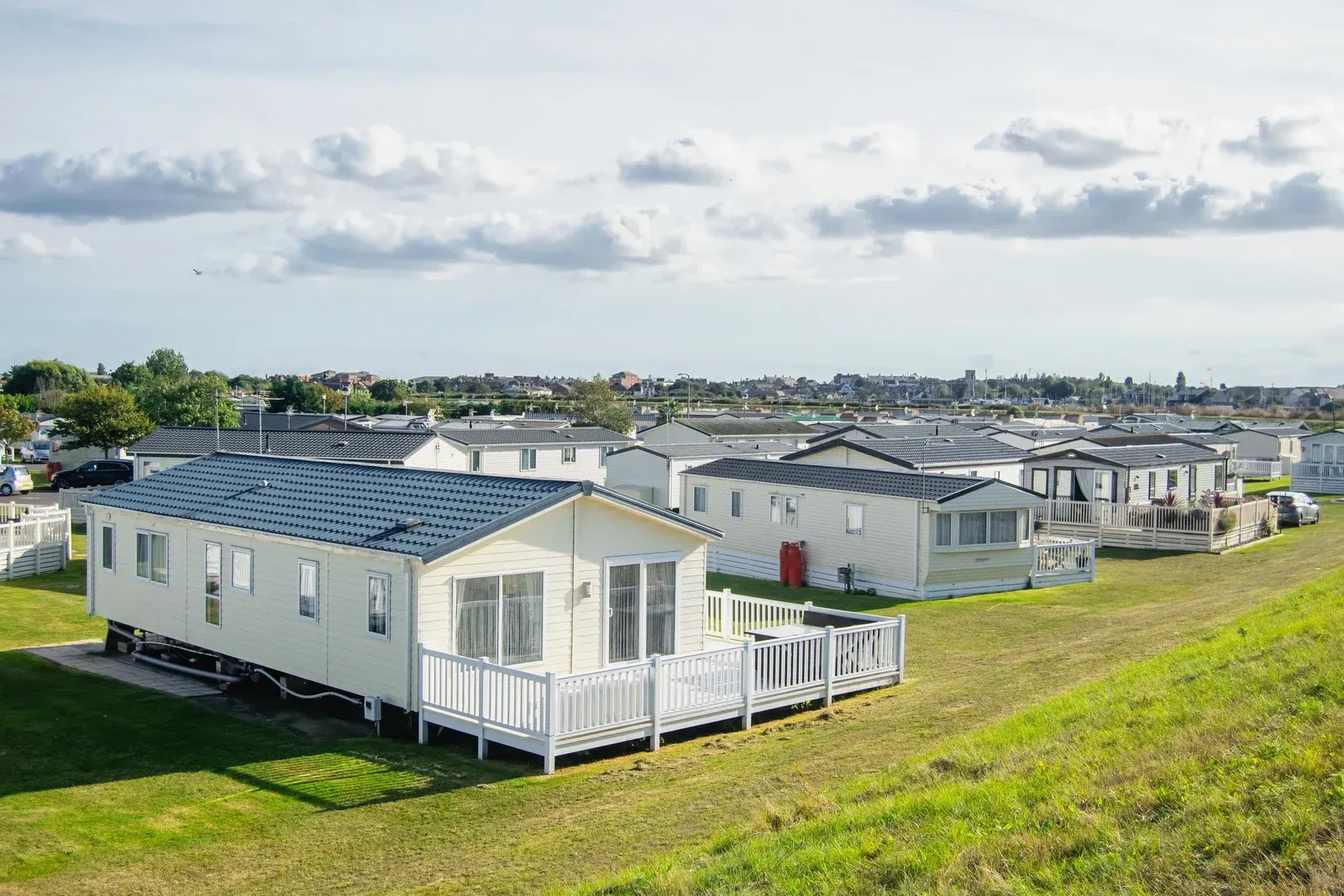 A row of cream-colored mobile homes with white decks arranged on a grassy site under a cloudy blue sky.