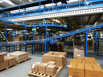 A warehouse floor featuring blue conveyor belt systems above and cardboard boxes stacked on wooden pallets below.
