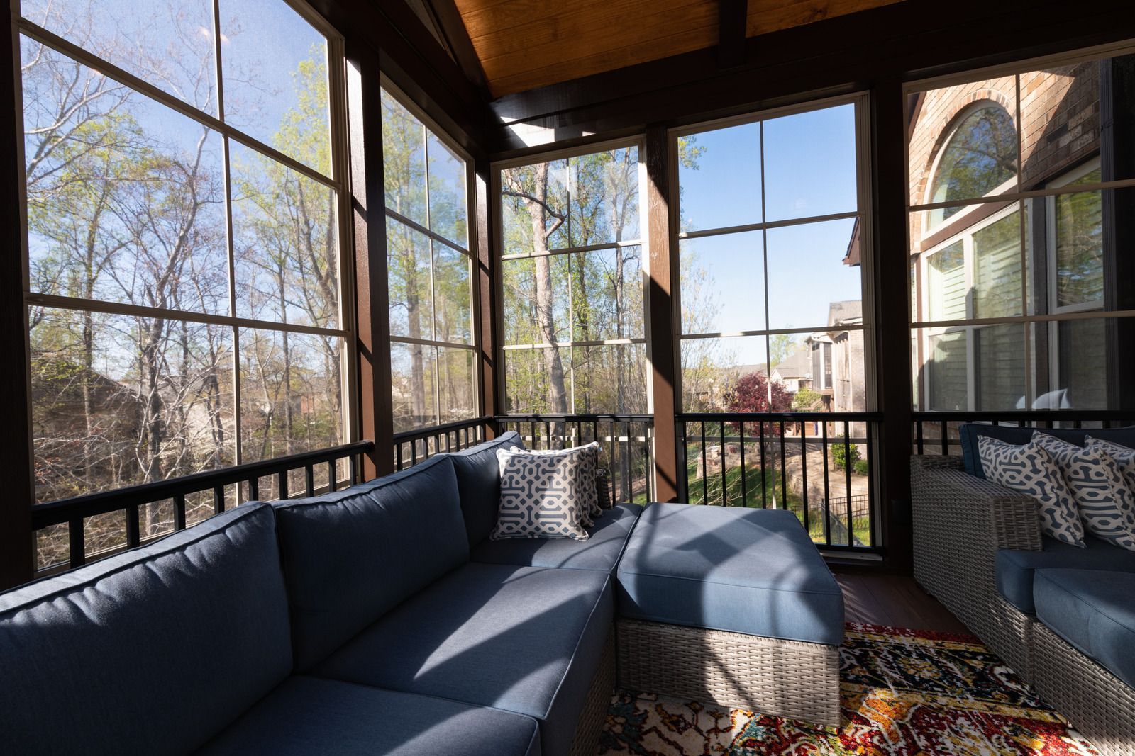 A cozy screened-in porch with blue sofa seating, dark wicker frames, and large windows overlooking trees and houses.