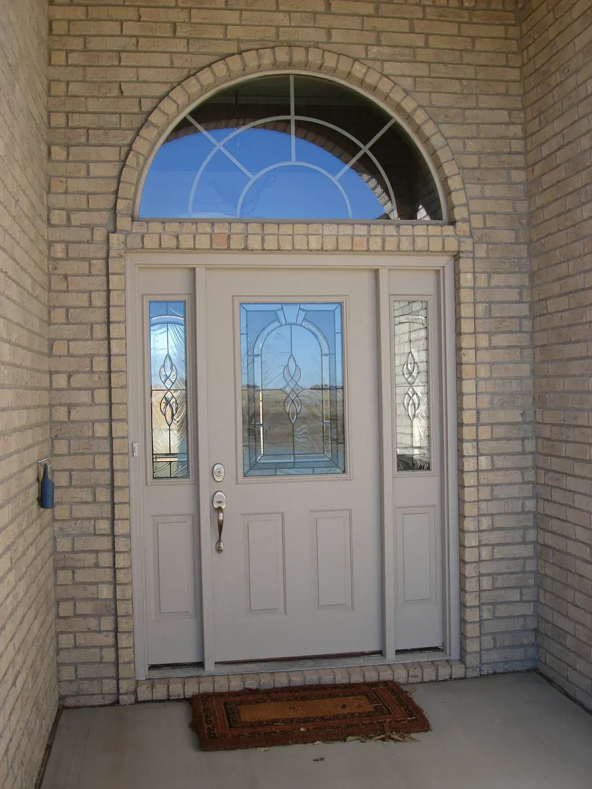 A light-colored front door with glass panels and a decorative arched transom window, set in a light brick wall.
