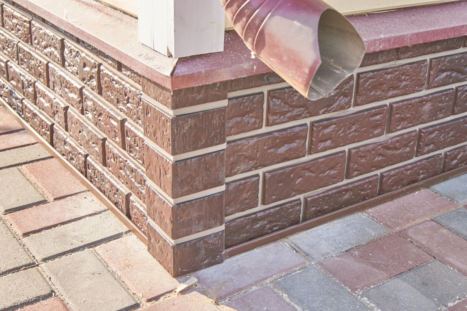 A dark brown brick house foundation with a corner pillar, a red downspout, and paved brick ground.