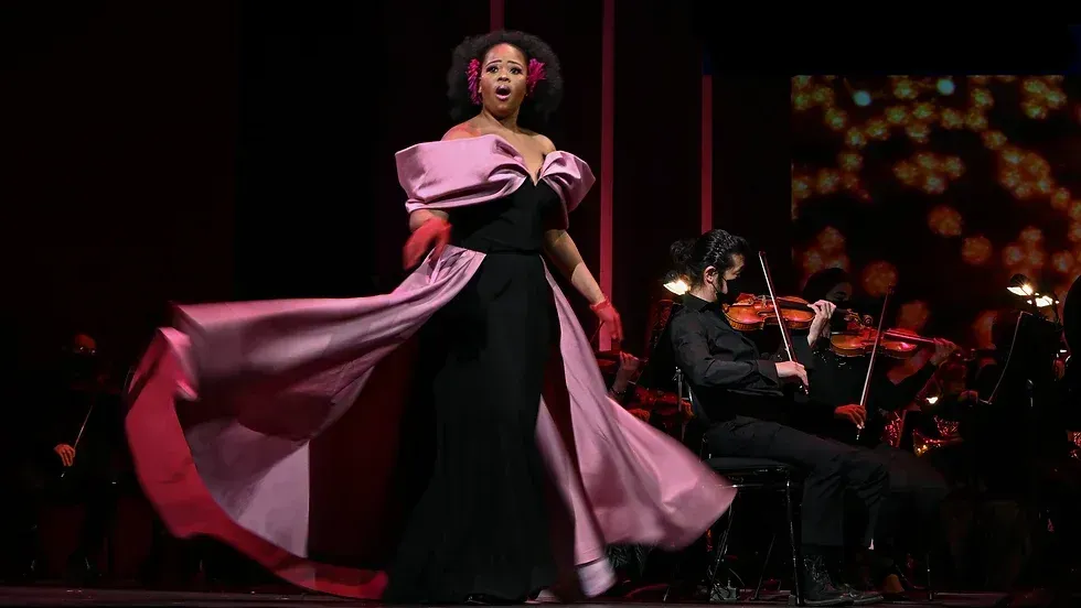 A woman in a pink dress is singing on a stage in front of an orchestra.