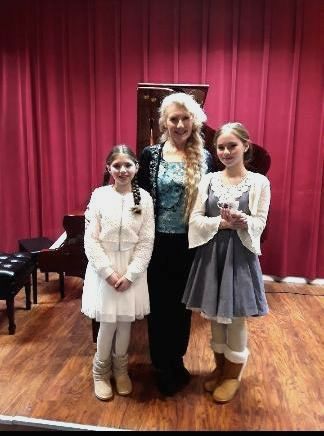 A woman is standing next to two young girls on a stage in front of a piano.