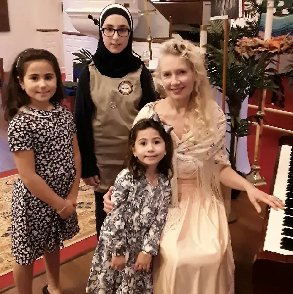 A woman is sitting in front of a piano with three girls standing around her