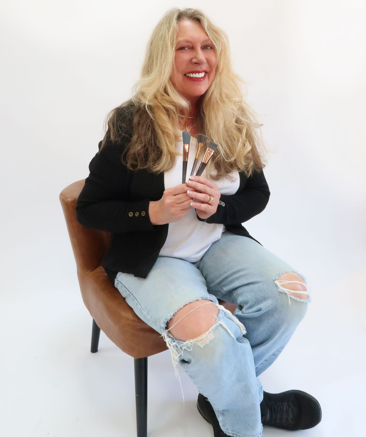 Connie holding makeup brushes, seated in a brown chair against a white background.