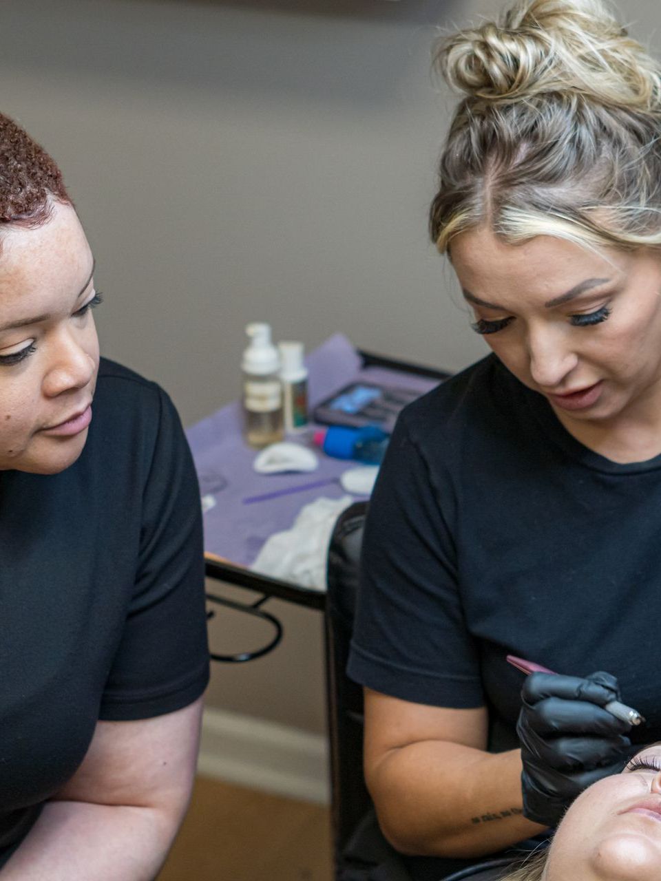 Two women are sitting at a table getting a tattoo on a woman 's face.