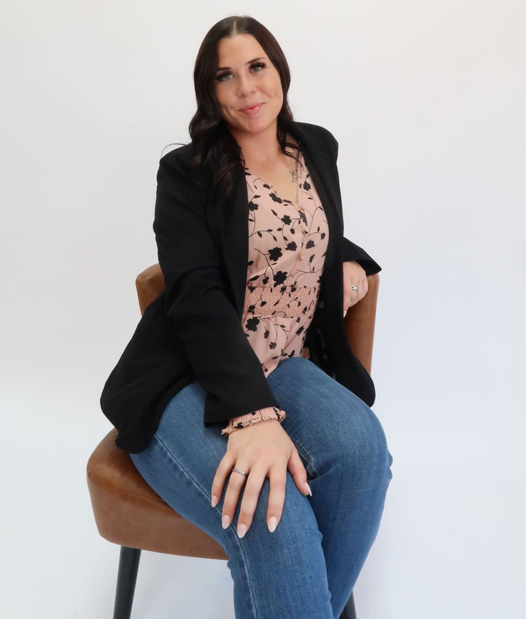 Woman seated in a chair, wearing a blazer, jeans, and floral top, against a white background.