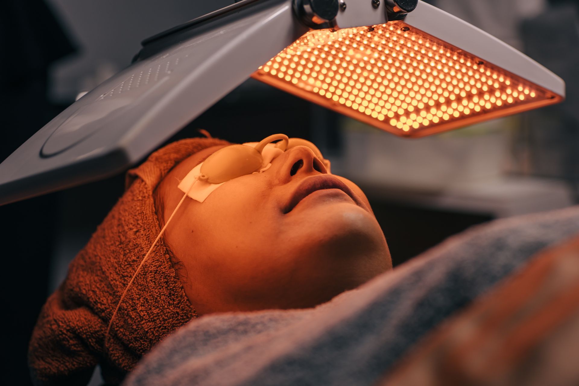 Person undergoing facial treatment under a red light device; wearing eye protection.