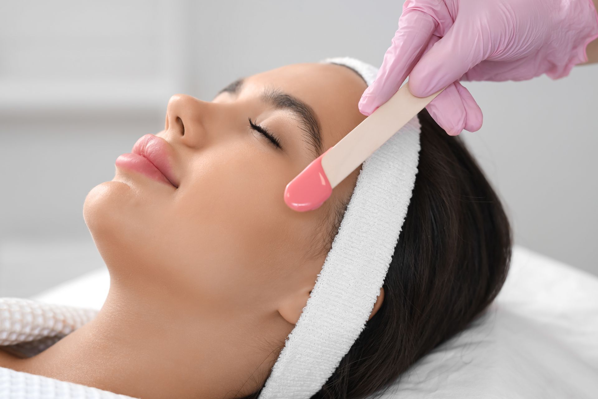 Woman getting facial waxing; pink wax applied to her cheek, in a salon setting.