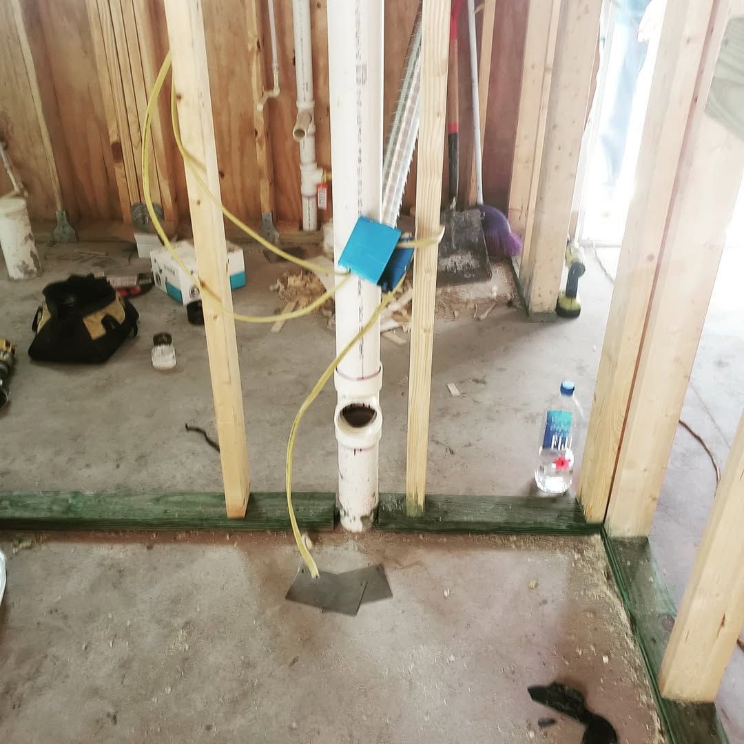 Interior construction of a bathroom. Framing surrounds a drain pipe in concrete floor. Wires and tools are scattered.