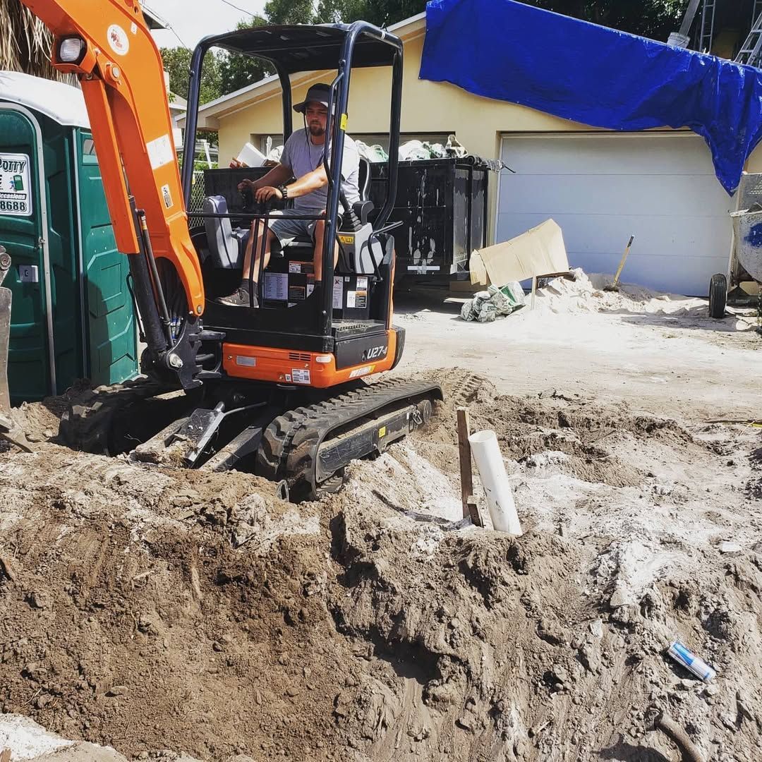 A person operating an orange excavator in a dirt pit, near a portable toilet and garage.