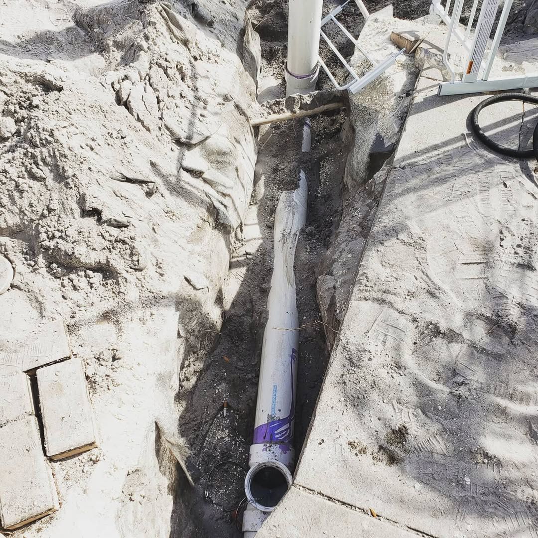 Trench dug in sand with exposed white PVC pipe. A partial view of a white railing is visible.