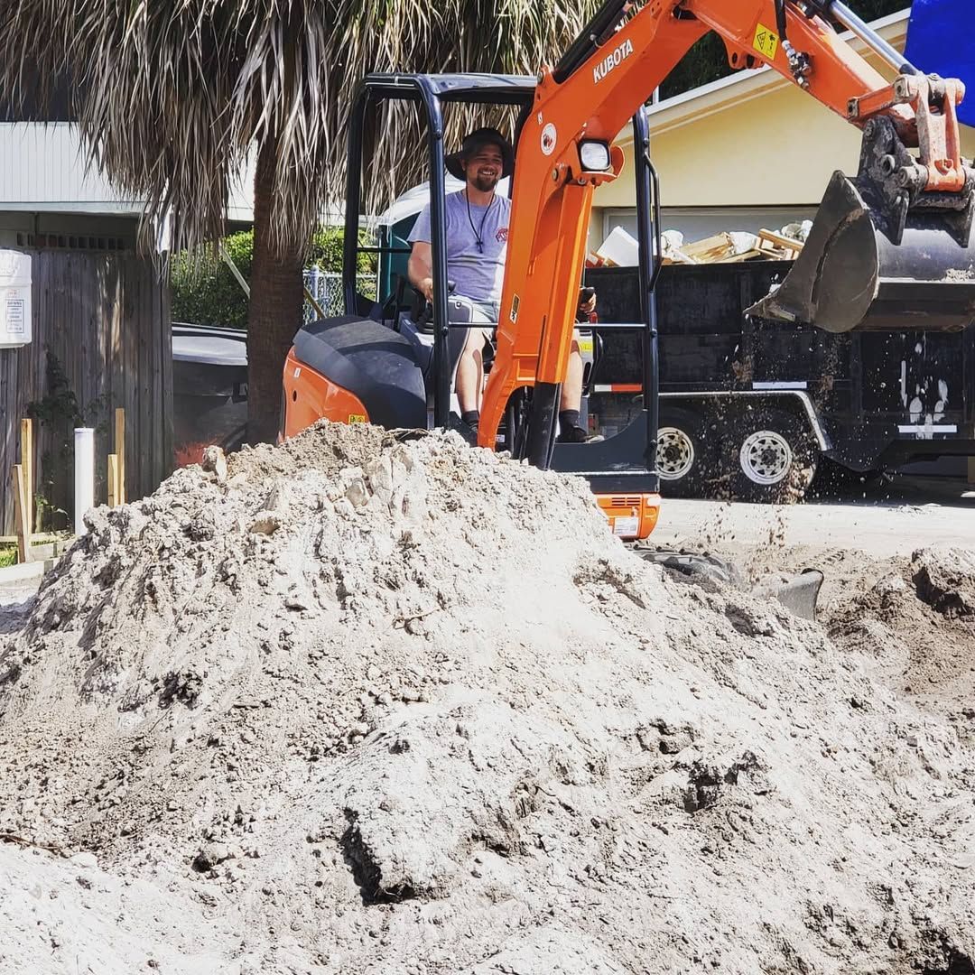 Person operating an orange excavator, moving dirt pile to a trailer, sunny outdoor setting.