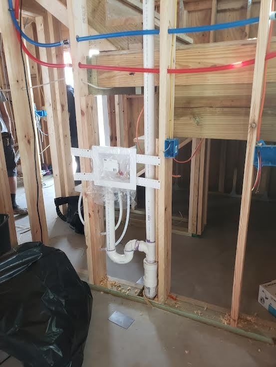 Framed wall with plumbing and electrical work in progress; white pipes, blue and red tubes, and blue electrical boxes.