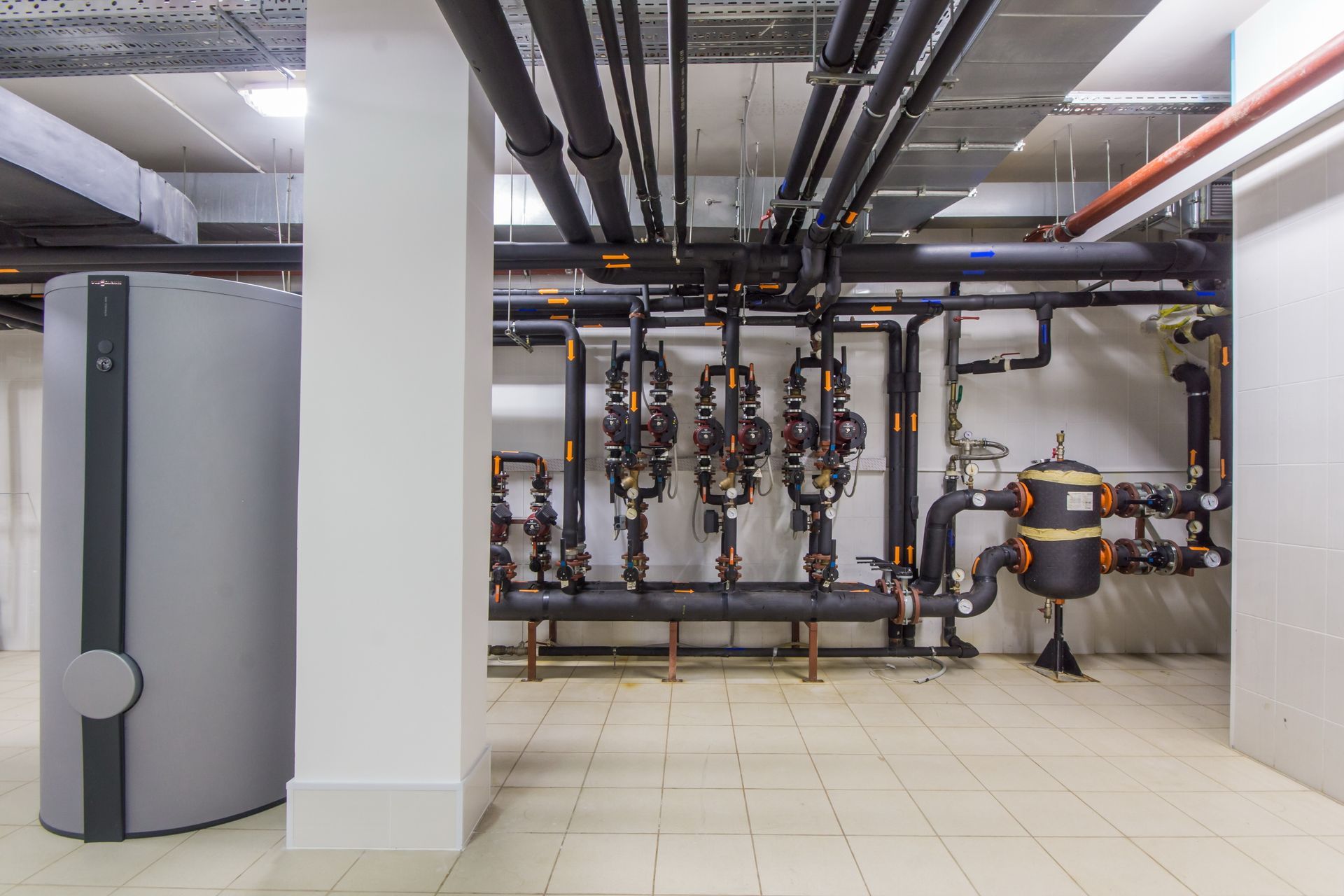 Mechanical room with pipes, tanks, and filtration system. Gray and black pipes against white walls and floor.