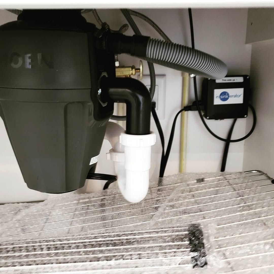 Garbage disposal under a kitchen sink. Grey disposal, white drain pipes, black electrical box, and a wire rack on the bottom.
