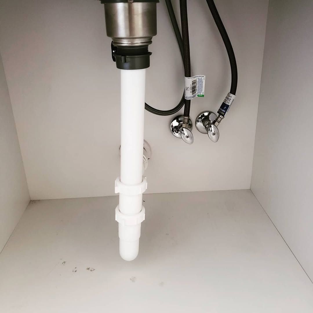 White under-sink cabinet with plumbing: white drain pipe, silver faucet and drain, and black water supply lines.