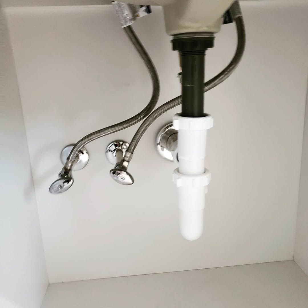 Underside of a white bathroom sink with plumbing visible. Flexible water supply lines connect to chrome valves. A white drain trap is present.