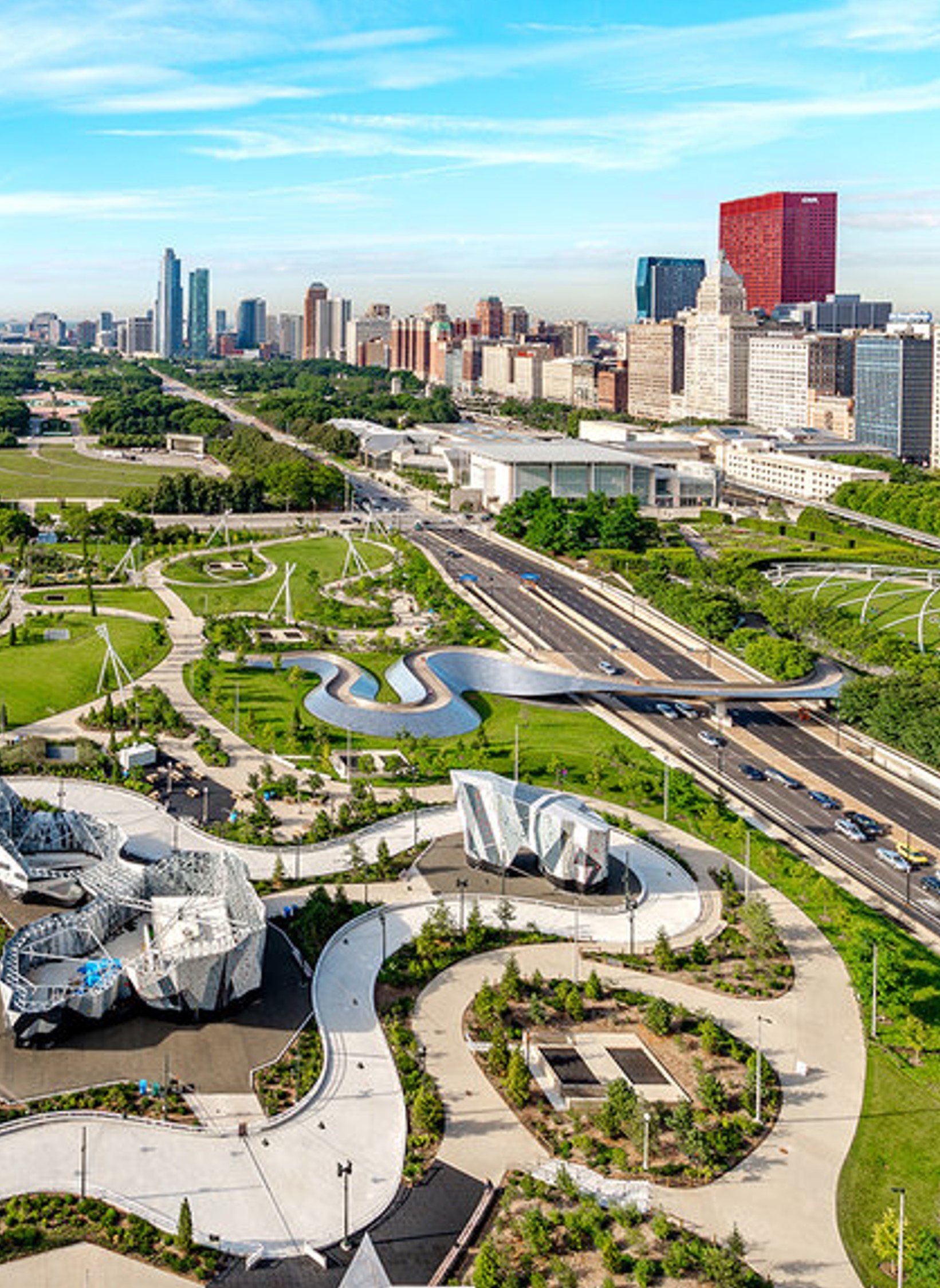 Aerial view of a modern park with winding paths, a highway, and a city skyline in the background.