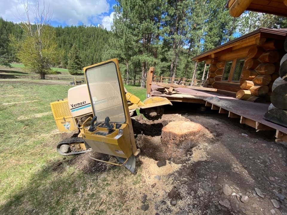 A yellow stump grinder removes a tree stump near a wooden deck and log cabin in a grassy area.