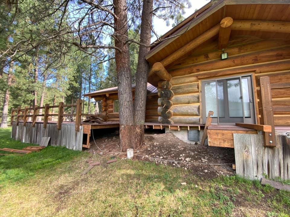 Log cabin with a wooden deck, a tree growing in the center. Sunny outdoor setting, green grass.