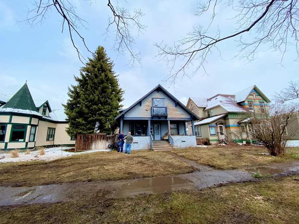 Muddy yard with several colorful historic houses. Two people stand on porch of a light blue house, possibly doing maintenance.