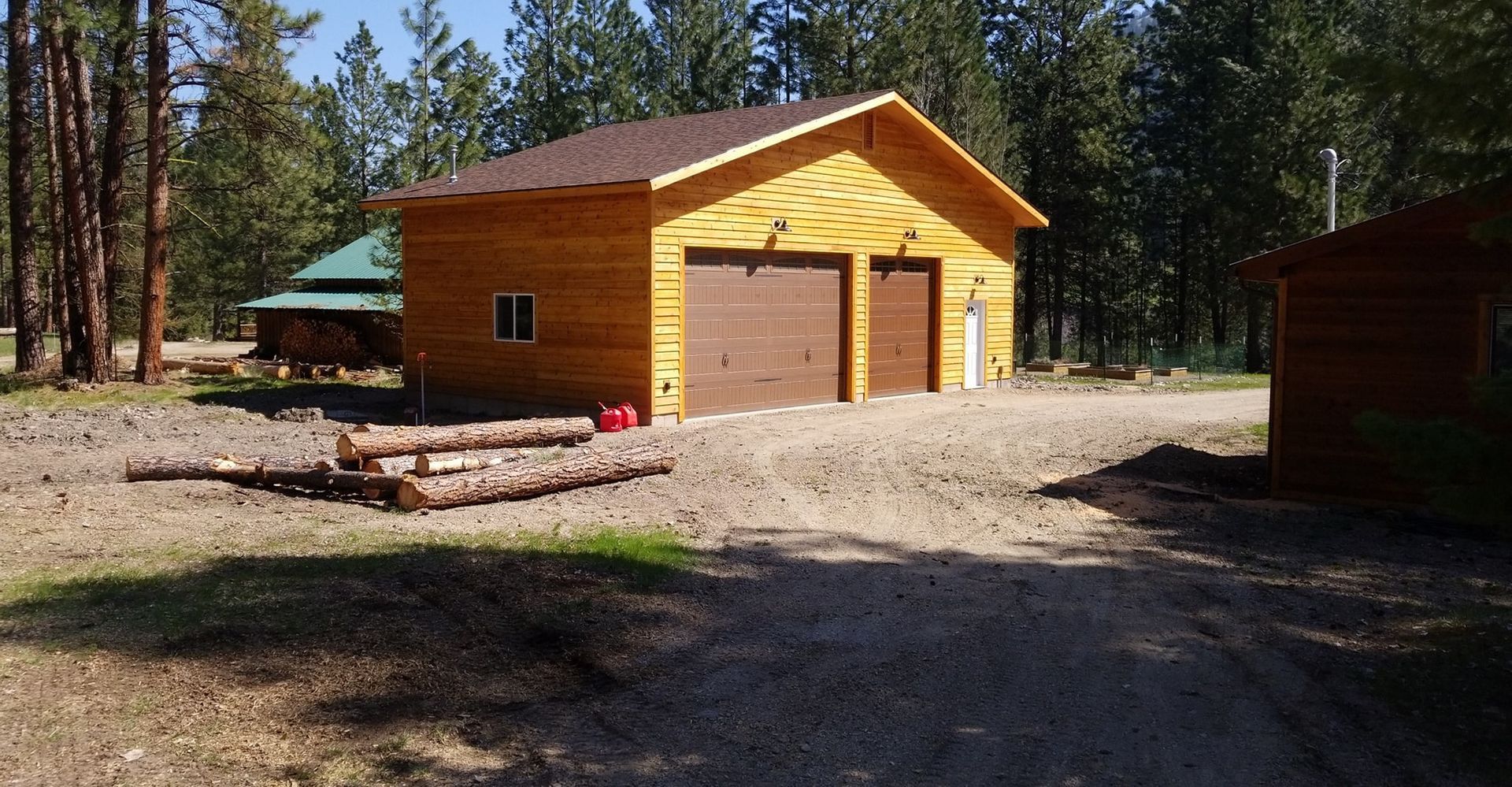 Yellow wooden garage with two brown doors on a gravel driveway, in a wooded area. A smaller building is visible in the background.