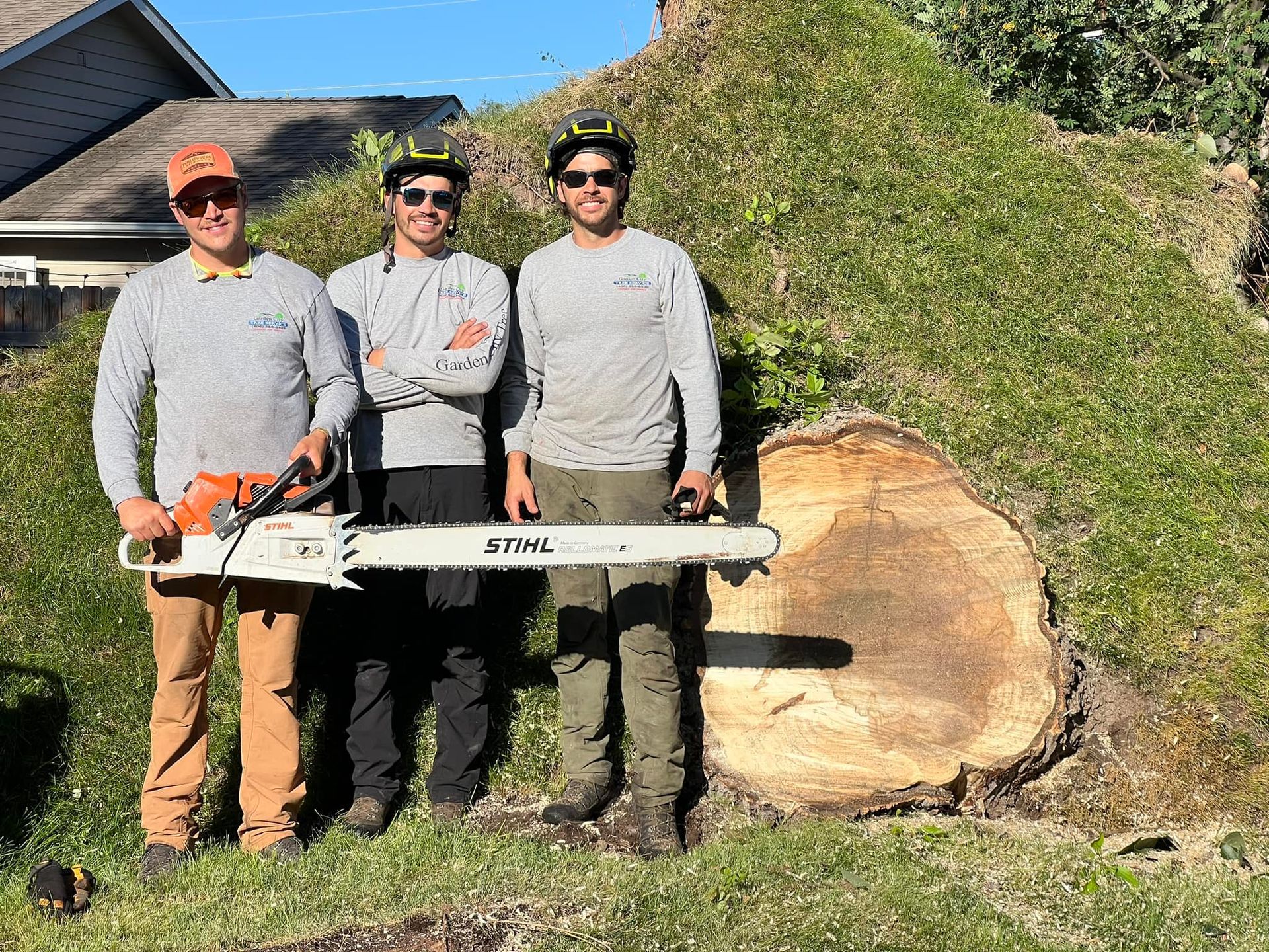Three arborists posing with a large chainsaw near a large cut tree trunk. They wear safety gear and casual clothing, smiling outdoors.