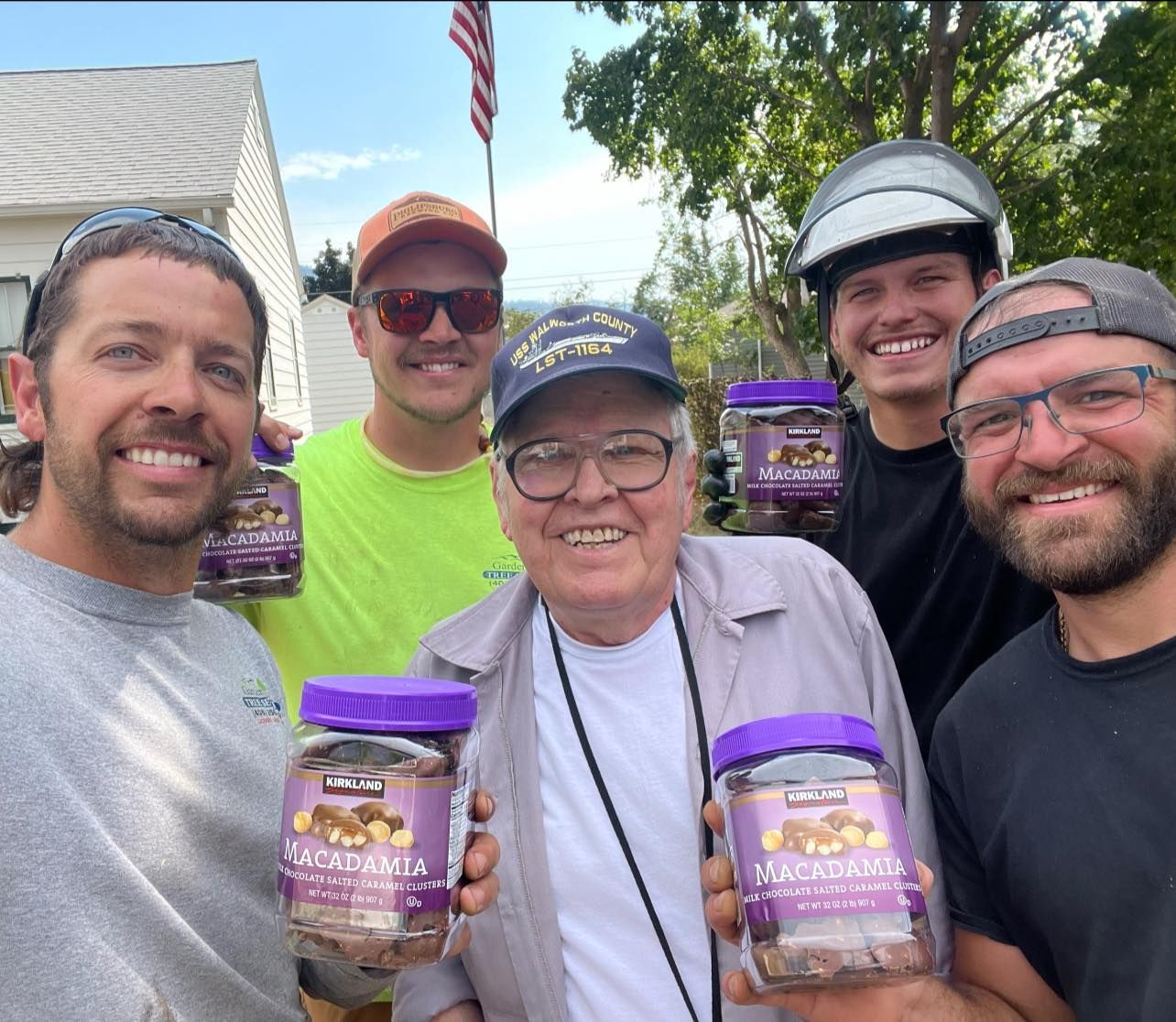A group of six people smiling and holding jars of macadamia nuts outside. One man is in the center, and the American flag is visible in the background.