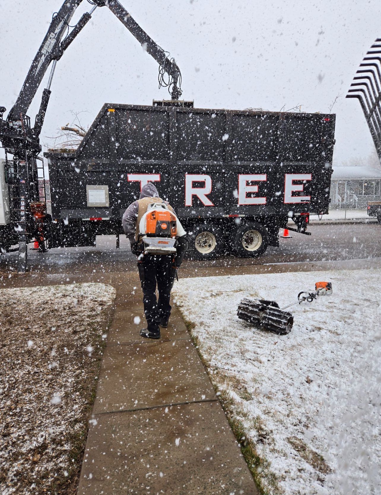 A tree service worker uses a leaf blower in the snow, in front of a truck that says 