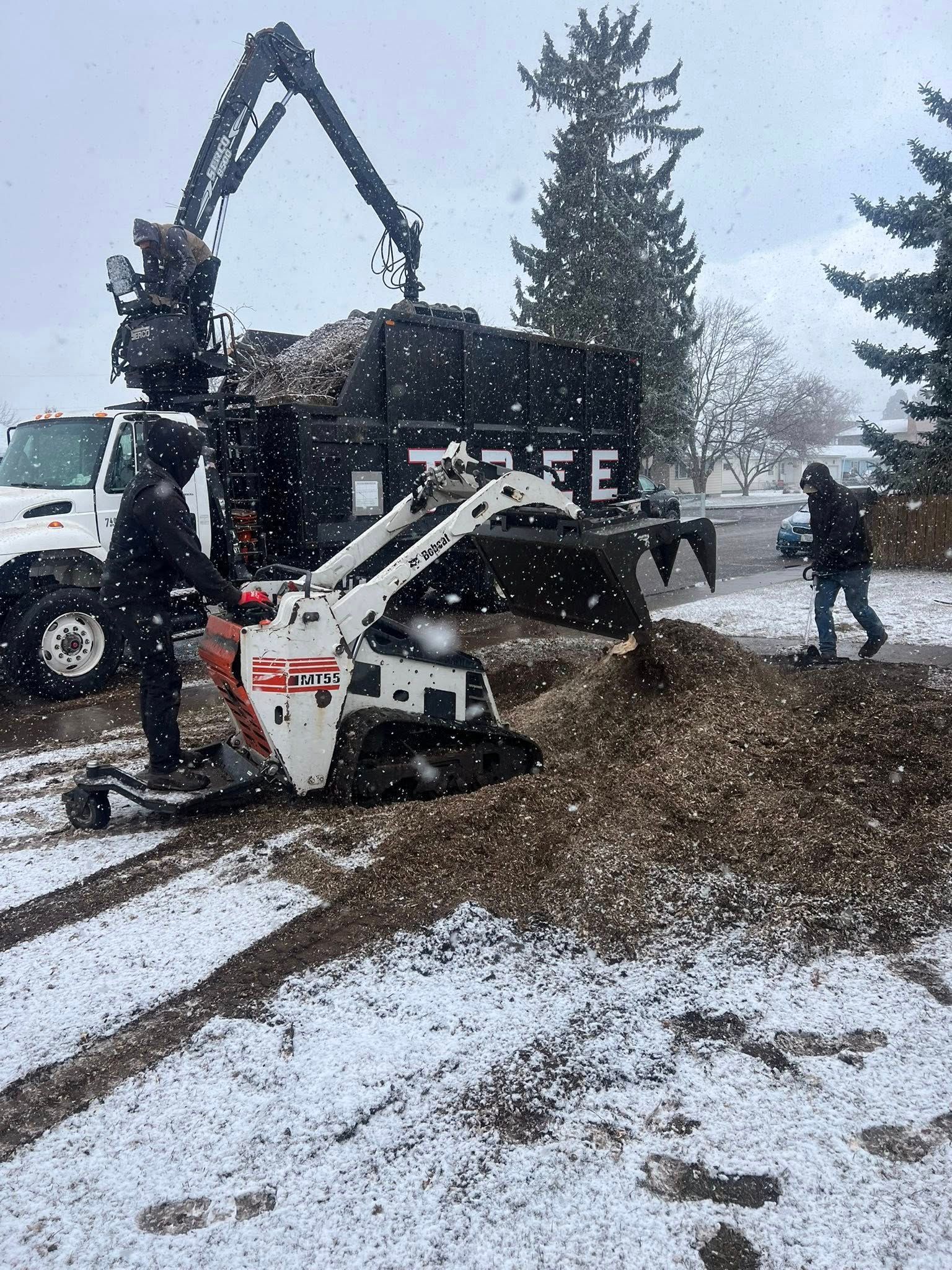 Two workers load gravel into a truck with a mini-excavator in a snowy outdoor setting.