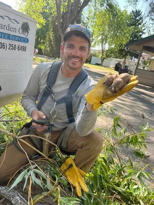 Man in work clothes smiles while holding baby animals in gloved hand near tree branches.  He is outside near a white vehicle.