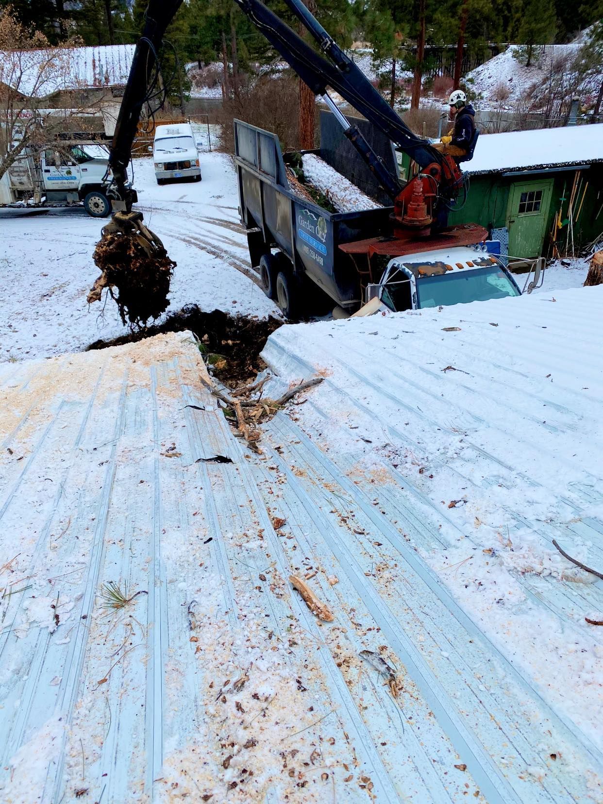 A truck with a crane emptying debris onto a snow-covered metal roof near a residential area.