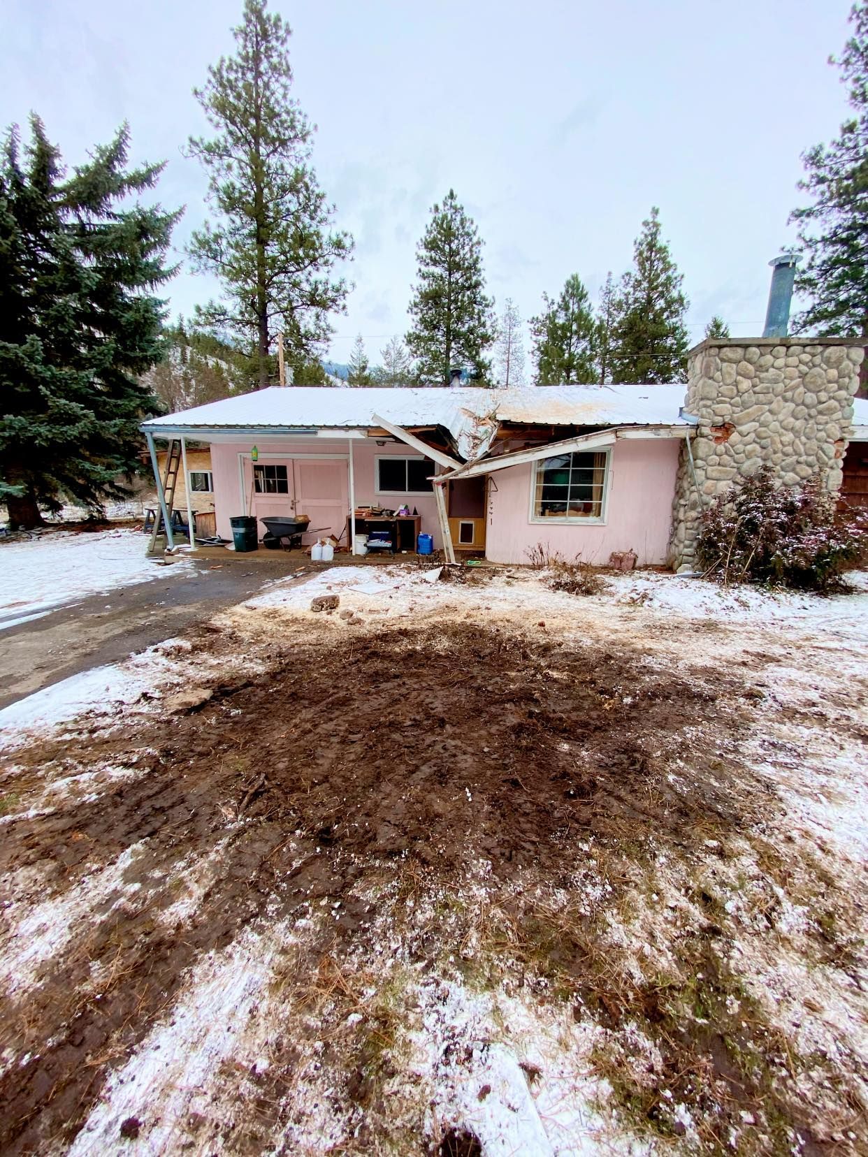 Pink house with significant roof damage, surrounded by snow-covered ground and bare trees.