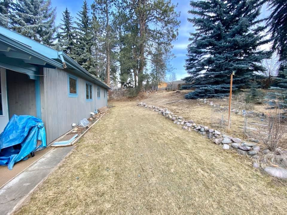 A long, narrow yard next to a light blue house, with a stone border along a path and tall trees in the background.