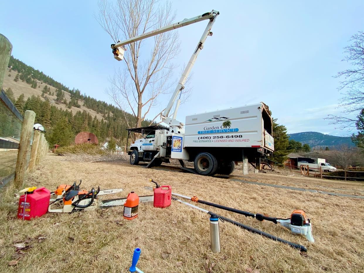 A tree service truck with an extended arm trimming a tree. Tools, including chainsaws and gas cans, are on the ground.