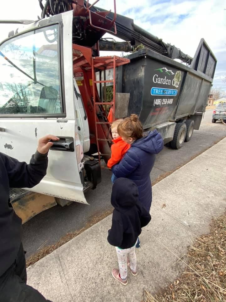 Woman holding a child stands near a large black dump truck. A man holds a phone, possibly taking a photo. The truck is on a street.