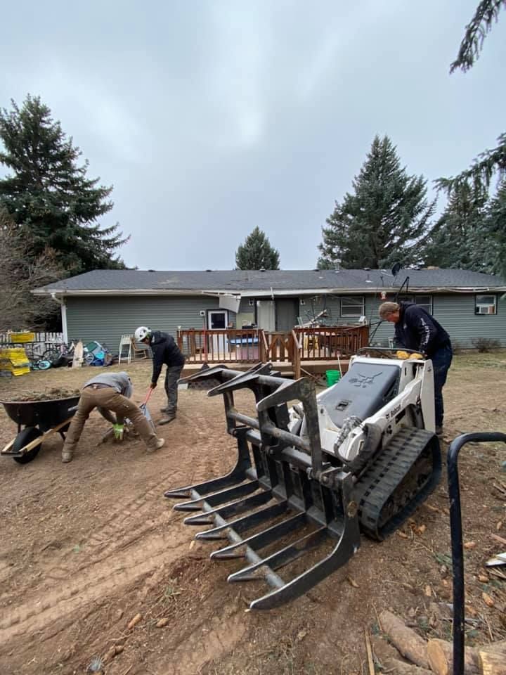 Four people working in a backyard with a small skid steer loader. They are moving dirt and debris, likely landscaping. Overcast day.