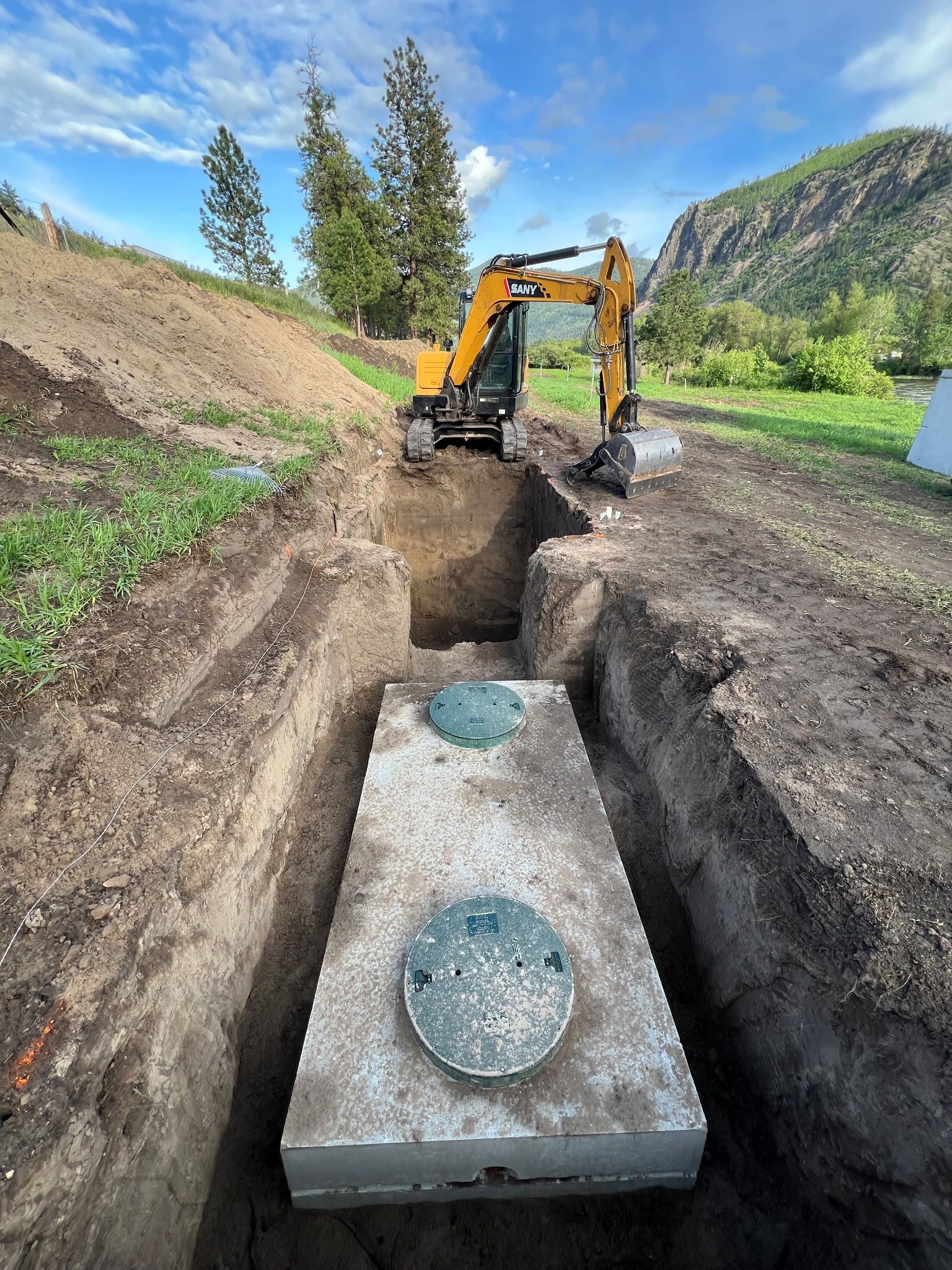 An excavator is working to bury a concrete septic tank in a deep trench. Green lid visible.  Mountains and blue sky in the background.