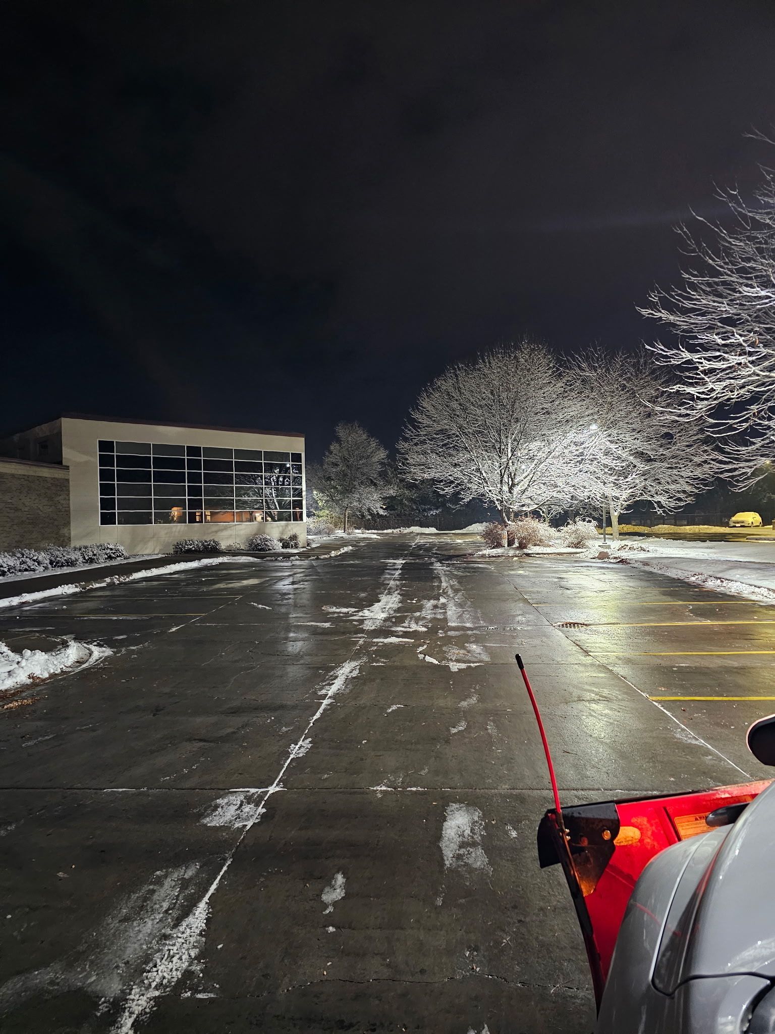 Snow-covered parking lot at night. A snowplow in foreground clears a path, with illuminated trees and building in the background.