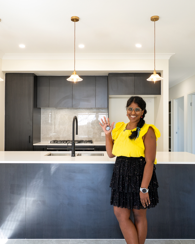 Woman in yellow top and black skirt in modern kitchen, smiling and waving.