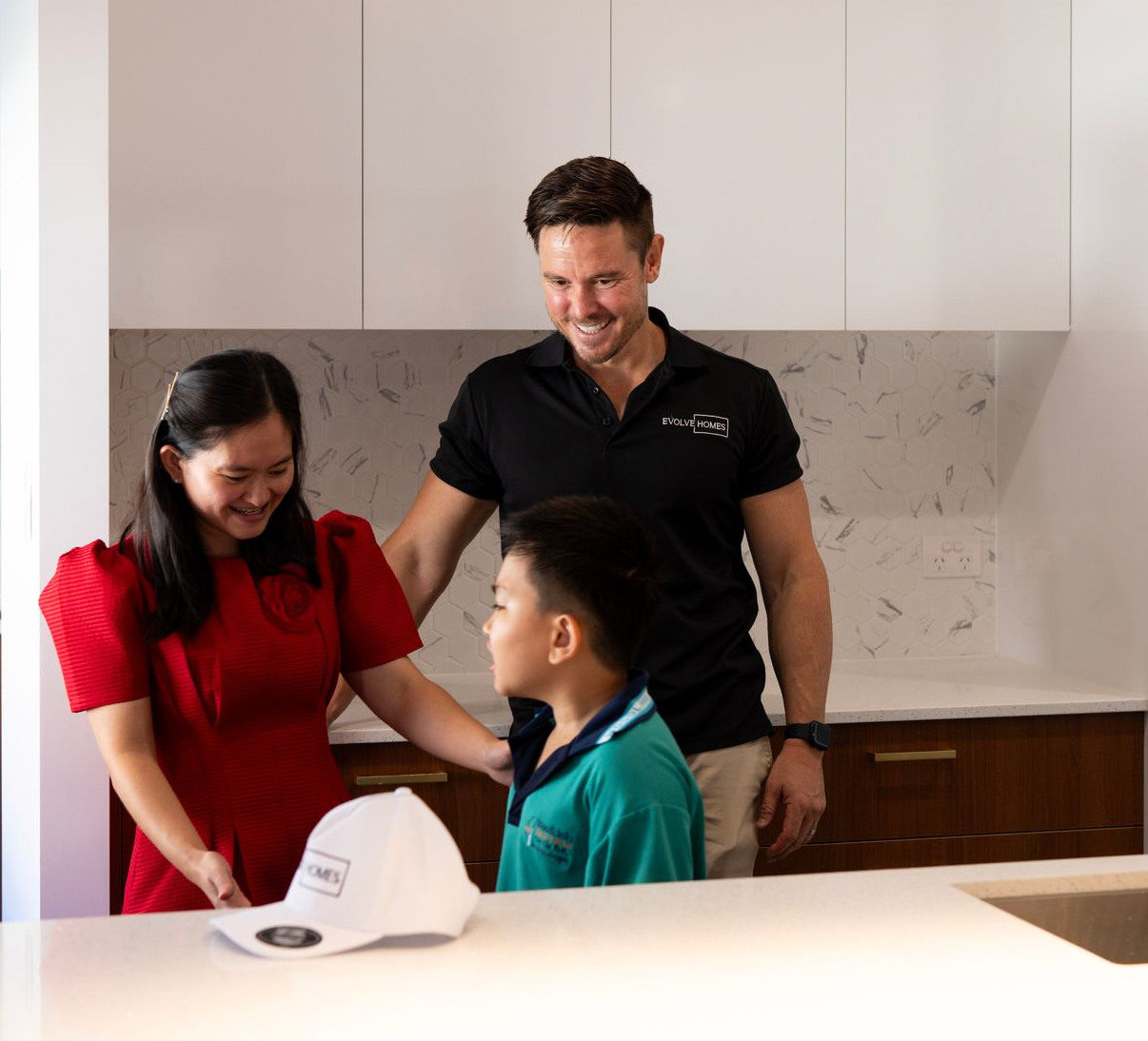 Family in a kitchen smiles at a white hat. Dark hair, red dress, black shirt with logo. Modern white and wood cabinets.