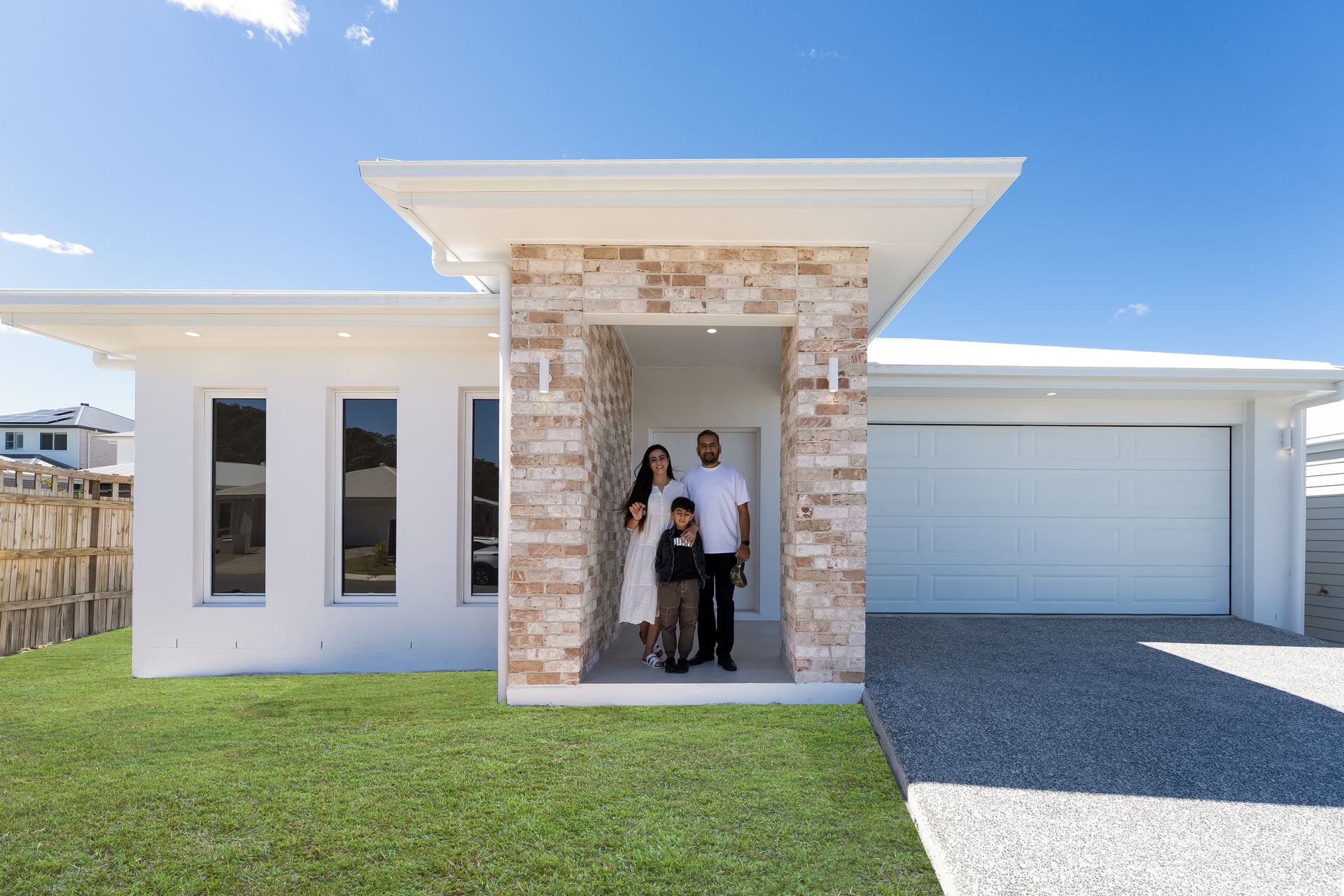 Family of three standing in front of a modern white house with stone accents, blue sky.